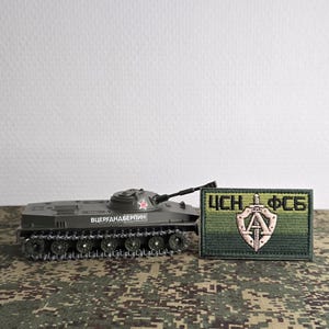 Może przedstawiać: Oliwkowozielony model miniaturowego czołgu z czerwoną gwiazdą i cyrylicą na boku, umieszczony obok prostokątnej haftowanej naszywki przedstawiającej tarczę, miecz oraz cyrylickie litery ЦСН i ФСБ wykonane zieloną i beżową nicią.
