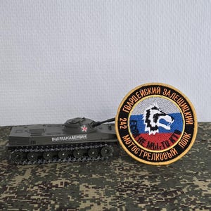 Puede incluir: Un modelo de tanque gris con una estrella roja en la torreta y el texto VTSERGDANDBERPIN en el lateral, colocado junto a un parche bordado circular con un gráfico de cabeza de lobo, la bandera rusa y texto cirílico en oro y negro.
