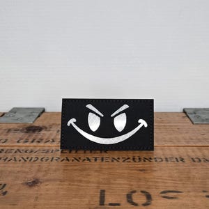 Può includere: Una toppa rettangolare nera con una grafica argentata riflettente che raffigura un ampio sorriso malizioso con occhi affilati e angolati. Il design presenta una sottile trama a nido d'ape negli elementi argentati e bordi cuciti.