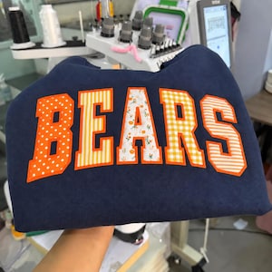 May include: A navy blue sweatshirt featuring the word BEARS in large, block-style letters. Each letter has a bright orange border and a unique pattern: white polka dots, vertical yellow stripes, orange floral print, yellow gingham, and horizontal stripes.