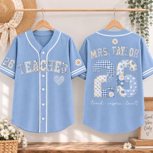 May include: A light blue button-down baseball jersey featuring white piping and patchwork-style lettering that reads TEACHER on the front and MRS. TAYLOR 26 on the back. The design includes blue and white gingham, floral patterns, and daisy motifs.
