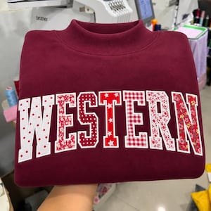 May include: A maroon sweatshirt featuring a mock neck collar and the word WESTERN appliqued across the chest. Each letter is crafted from a different fabric pattern, including white polka dots, red floral, red gingham, and red star prints.