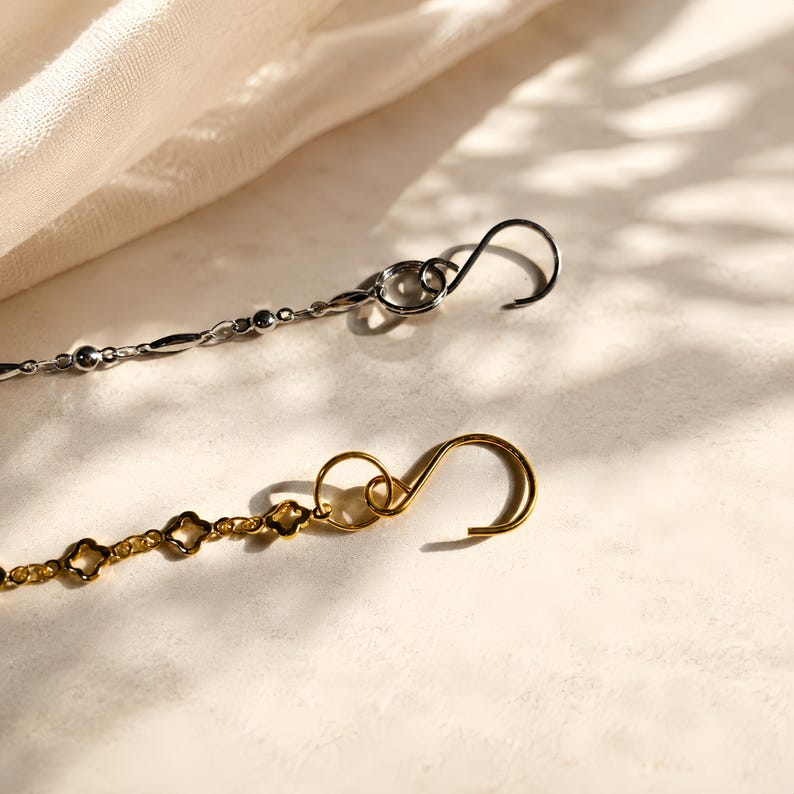 Puede incluir: Dos cadenas de metal, una plateada y otra dorada, cada una con un gancho en forma de S en el extremo. La cadena plateada tiene peque&ntilde;as cuentas esf&eacute;ricas y eslabones alargados, mientras que la dorada consiste en una serie de peque&ntilde;os eslabones abiertos en forma de tr&eacute;bol.