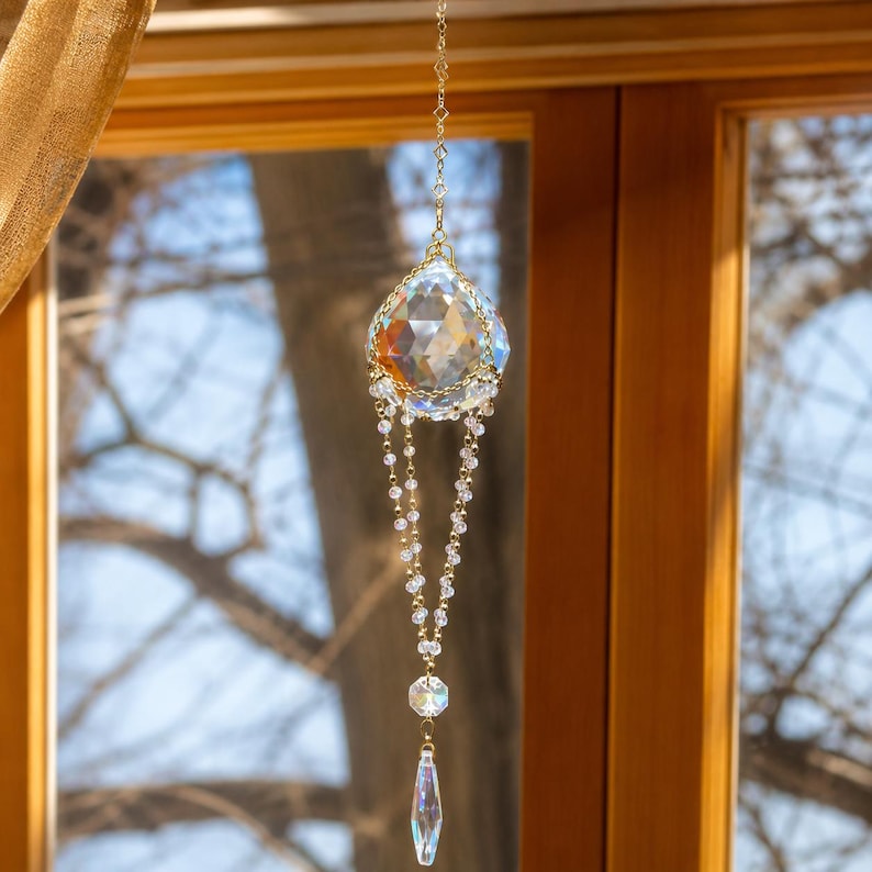 Puede incluir: Un atrapasoles de cristal colgante que presenta una gran esfera de vidrio transparente multifacetado suspendida por una cadena de metal dorado. Debajo de la esfera, dos delicadas cadenas de cuentas cuelgan hacia abajo, conectadas a un peque&ntilde;o cristal hexagonal y un prisma en forma de l&aacute;grima.