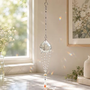 Puede incluir: Un atrapasoles de cristal colgante con una gran esfera de vidrio transparente facetado suspendida de una cadena de metal plateado. Varias hebras delicadas de cuentas cuelgan de la esfera, terminando en un prisma de gota de vidrio claro.