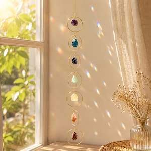 May include: A vertical hanging ornament featuring seven gold-toned metal rings linked together in a chain. Each ring contains a single raw, uncut gemstone in varying colors including purple, blue, green, yellow, orange, and deep red.
