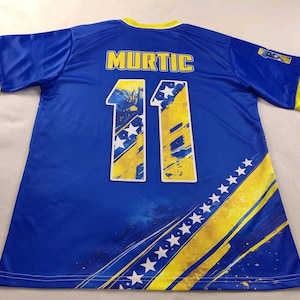 Puede incluir: Una camiseta de f&uacute;tbol azul con el nombre "MURTIC" en la espalda y el n&uacute;mero "11" en amarillo y blanco. La camiseta tiene detalles amarillos y blancos en las mangas y los laterales.