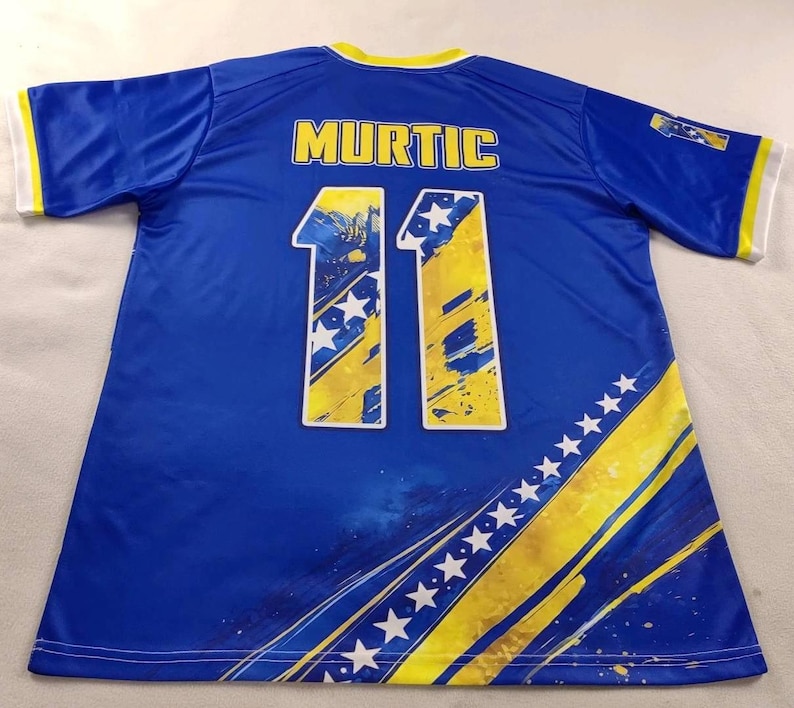 Puede incluir: Una camiseta de f&uacute;tbol azul con el nombre "MURTIC" en la espalda y el n&uacute;mero "11" en amarillo y blanco. La camiseta tiene detalles amarillos y blancos en las mangas y los laterales.