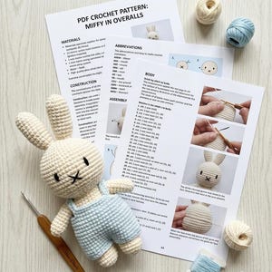 Könnte beinhalten: Eine gehäkelte cremefarbene Kaninchenpuppe mit hellblauer Latzhose, platziert neben einer ausgedruckten PDF-Häkelanleitung mit dem Titel Miffy in Overalls, einer Holzhäkelnadel und mehreren kleinen Knäueln aus cremefarbenem und hellblauem Garn.