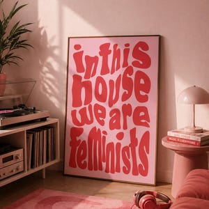 Könnte beinhalten: Ein rechteckiges gerahmtes Wandbild mit hellrosa Hintergrund und fett gedrucktem, wellenförmigem rotem Text, der "in this house we are feminists" lautet. Das Poster befindet sich in einem dünnen Rahmen und steht aufrecht auf dem Boden.