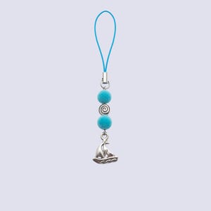 Peut inclure: Une breloque pour téléphone composée d'une boucle bleu vif, de deux sphères turquoise duveteuses, d'une perle en métal argenté avec un motif en spirale et d'un pendentif en métal argenté en forme de voilier suspendu à la base.