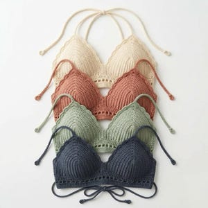 Può includere: Una pila di quattro top all'americana in uncinetto nei colori crema, terracotta, verde salvia e blu navy scuro. Ogni top presenta un design a coppe triangolari con bordo smerlato, una delicata fascia traforata alla base e sottili laccetti.