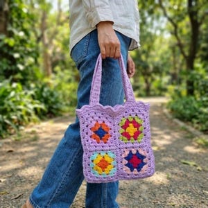 May include: A lavender crochet tote bag featuring four square granny squares with colorful centers in shades of red, yellow, blue, and green. The bag has two matching lavender handles and a scalloped top edge, constructed from yarn in a textured pattern.