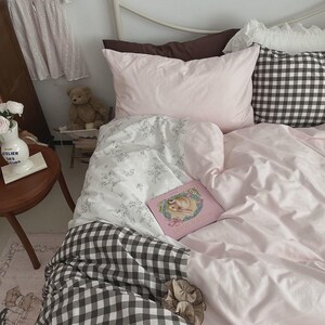 Puede incluir: Una cama con ropa de cama de color rosa claro con estampado floral blanco, una manta de cuadros vichy en blanco y negro, y un libro rosa titulado Maiden Diary. Un jarrón blanco con flores blancas, un oso de peluche y una prenda blanca.