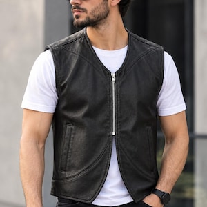 May include: A black leather vest featuring a central silver zipper closure, a V-neckline, and two vertical side pockets. The vest is worn over a plain white short-sleeved t-shirt, showcasing a clean, sleeveless design with structured seams.