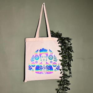 Könnte beinhalten: Eine hellrosa Tragetasche mit einer zentralen Grafik einer Person mit Blumenkranz und gemusterter Kleidung. Das Design ist in einem lebendigen, irisierenden Regenbogen-Farbverlauf gehalten, der einen reflektierenden Effekt erzeugt.
