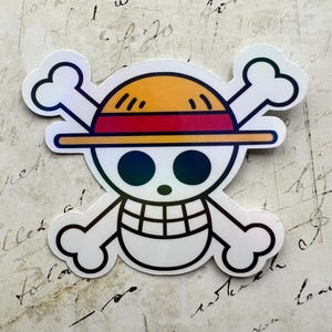 Könnte beinhalten: Ein gestanzter Aufkleber mit dem Jolly Roger Emblem aus One Piece. Das Design zeigt einen weißen Totenkopf mit einem gelben Strohhut mit rotem Band, der über zwei gekreuzten weißen Knochen liegt. Der Totenkopf hat große, dunkle Augenhöhlen mit Farbverlauf.
