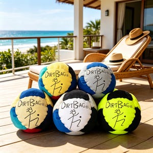 Puede incluir: Seis pelotas Dirtbag multicolores están dispuestas sobre una terraza de madera. Cada pelota tiene una combinación de colores diferente, incluyendo azul, amarillo y negro, con el texto "DIRTBAG" y un logotipo de figura de palo. Se ve una escena de playa al fondo.