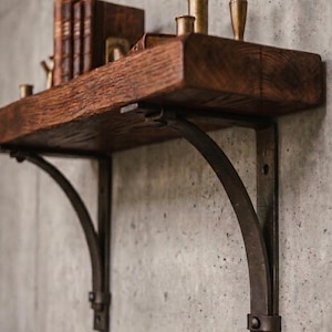 Peut inclure: Une étagère épaisse en bois brun foncé avec un bord naturel brut, soutenue par deux supports métalliques sombres incurvés. L'étagère porte trois livres verticaux et trois petits bougeoirs cylindriques métalliques de hauteurs variées.