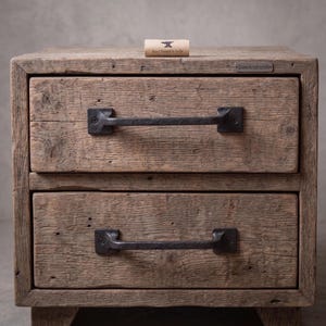 Op de afbeelding: Een houten nachtkastje met twee lades, elk voorzien van een donkere, handgesmede ijzeren handgreep. Het hout heeft een verweerde, getextureerde afwerking. Een label op de bovenste lade vermeldt SteelAndIronIN en Hand Forged in India.