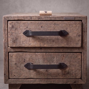 May include: A rustic wooden nightstand with two drawers, each featuring a matte black metal handle secured with screws. The wood has a weathered, textured grain. A small label on the top right reads SteelAndIronIN, and a tag on top says Hand Forged in India.
