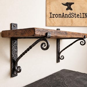 Pode incluir: Uma prateleira de parede com uma tábua grossa de madeira recuperada castanho-escura, suportada por dois suportes de ferro forjado preto com textura martelada, braços de metal torcido e extremidades decorativas em espiral. Uma placa acima diz IronAndStelIN.