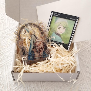 May include: A white cardboard box filled with shredded paper containing an acrylic standee featuring an illustrated character in a blue cloak and a square photo print framed by a black film strip border with the text STAX 15 ISO 800 F5.6 1/1000.