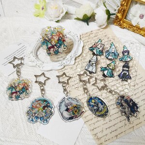May include: A collection of acrylic merchandise featuring anime-style character illustrations. The set includes five star-clasp keychains, one acrylic standee, and six small pins, all depicting figures in pointed hats and robes with colorful, detailed artwork.
