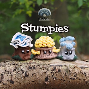 Pode incluir: Três figuras Stumpies de crochê com rostos sorridentes. A figura da esquerda tem uma base castanha e um topo azul com folhos. A do meio tem uma base castanha clara e um topo amarelo texturado. A da direita tem uma base cinzenta e um topo de cogumelo azul.