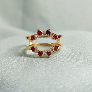 May include: A gold-colored ring with a double band design. The top band features a series of small, round, red gemstones alternating with clear, round gemstones. The ring is set against a soft, light blue background.