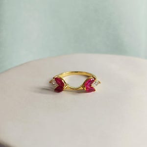 Könnte beinhalten: Ein goldfarbener Ring mit geschwungenem Design, der auf jeder Seite drei rote Edelsteine und an jedem Ende einen weißen Edelstein aufweist. Der Ring liegt auf einer weißen Oberfläche mit hellblauem Hintergrund.