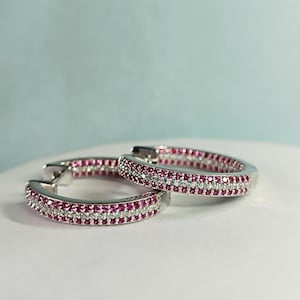May include: A pair of silver hoop earrings featuring three parallel rows of stones. The outer rows are set with small, vibrant pink circular stones, while the center row consists of clear, sparkling white stones, creating a multi-layered design.