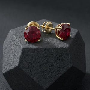May include: Gold-coloured stud earrings featuring large, round, deep red gemstones. The earrings are presented on a dark grey, geometric-shaped surface, highlighting their colour and design. The earrings have a classic, elegant style.
