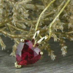 Könnte beinhalten: Eine Goldkette mit einem facettierten, herzförmigen roten Edelsteinanhänger. Der Anhänger wird von einer goldenen Öse gehalten, die an einer feinen Goldkette befestigt ist. Der tiefrote Stein hat einen Brillantschliff, der das Licht reflektiert.