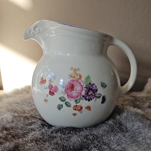 Może przedstawiać: Kremowy dzbanek ceramiczny z kwiatowym wzorem przedstawiającym różowe róże, fioletowe chryzantemy oraz małe pomarańczowe i żółte kwiatki. Dzbanek ma zaokrąglony korpus, zakrzywiony uchwyt oraz dzióbek z cienką niebieską obwódką na górnej krawędzi.