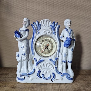 Op de afbeelding: Een witte keramische schoorsteenklok met blauwe handgeschilderde bloem- en krulmotieven. De wijzerplaat bevindt zich tussen twee staande figuren die elk een blauwe kan vasthouden. De wijzerplaat toont zwarte cijfers en een wereldbol.
