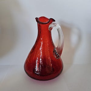 Puede incluir: Una jarra de vidrio rojo con un asa de vidrio transparente. La jarra tiene una superficie texturizada y un borde con volantes. El asa es curva y está unida al costado de la jarra. Probablemente se utiliza para servir bebidas.