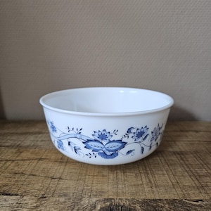 Op de afbeelding: Een witte glazen kom met een herhalend blauw bloemenpatroon aan de buitenzijde. Het ontwerp bestaat uit gestileerde blauwe bloemen, bladeren en dunne ranken die de omtrek van de kom omcirkelen, voorzien van een gladde, ronde rand en basis.