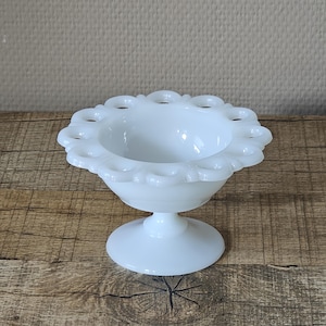 Puede incluir: Un cuenco blanco con pedestal de estilo vintage con un borde festoneado y una base redonda. El cuenco tiene un delicado diseño calado alrededor del borde superior. El cuenco está hecho de vidrio blanco opaco y se encuentra sobre una superficie de madera.