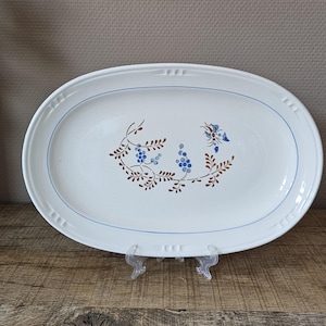 May include: An oval white ceramic serving platter featuring a central hand-painted botanical motif with brown leaves and blue berries. The rim is decorated with a thin blue circular line and embossed rectangular accents along the outer edge.