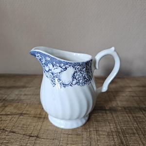 Op de afbeelding: Een witte keramische melkkan met een blauw transferpatroon van diverse vruchten en bladeren rond de bovenrand. De kan heeft een geribbelde body, een gebogen handvat en een schenktuit, wat een klassiek ontwerp laat zien.