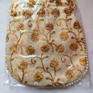Può includere: Una pochette con coulisse color crema caratterizzata da ricami in filo dorato con un motivo a tralci floreali. I bordi sono impreziositi da una fila di piccole perle bianche e la borsa è racchiusa in una custodia protettiva in plastica.