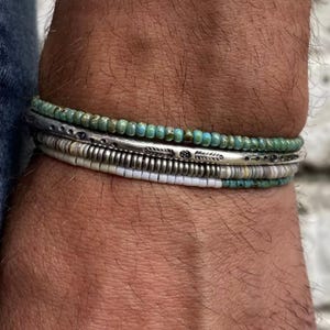Può includere: Una pila di quattro braccialetti composta da un filo superiore di piccole perline color turchese, una fascia centrale in metallo argentato con motivi a foglie e punti incisi, una fascia metallica a coste e un filo inferiore di perline bianche.