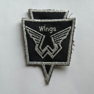 Könnte beinhalten: Ein schwarzer gestickter Aufnäher in Schildform mit silbernem Rand. Das Design zeigt ein stilisiertes silbernes W in Flügelform, wobei das Wort Wings in silbernem Garn über dem zentralen grafischen Element eingestickt ist.