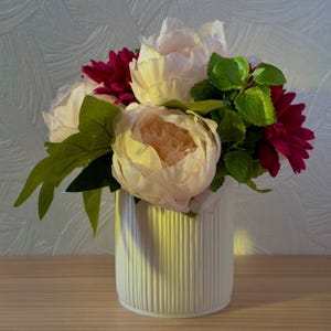 Può includere: Un'arrangiamento floreale in un vaso bianco a coste. Comprende peonie color crema, dalie rosso scuro e foglie verdi. La composizione crea un'atmosfera decorativa per la casa.