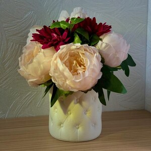 Può includere: Un'arrangiamento floreale con grandi peonie color crema e dalie rosso scuro, accentuato da foglie verdi. I fiori sono disposti in un vaso color crema trapuntato, creando un'esposizione morbida ed elegante. Il vaso è posto su una superficie di legno.