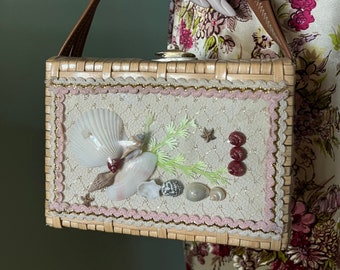 Könnte beinhalten: Eine geflochtene Strohtasche mit einem braunen Ledergriff. Die Vorderseite der Tasche ist mit Muscheln, Perlen und einer Spitzenborte verziert.
