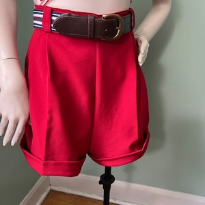 Könnte beinhalten: Rote, hoch taillierte Shorts mit einem braunen Ledergürtel und einem gestreiften Stoffgürtel. Die Shorts haben einen umgeschlagenen Saum.