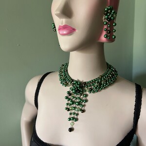 Può includere: Un set di collana e orecchini con perline verdi. La collana presenta un design a cascata con una parte superiore in stile choker. Gli orecchini sono di stile pendente con un gruppo di perline nella parte superiore.