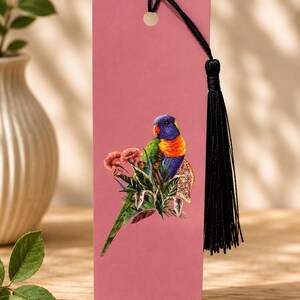Puede incluir: Un marcapáginas rectangular de color rosa empolvado con una vibrante ilustración de un lori arcoíris posado entre hojas verdes y flores rosas. Un cordón negro con una borla negra a juego está sujeto en la parte superior. El texto Original By Sam está impreso en la parte inferior.